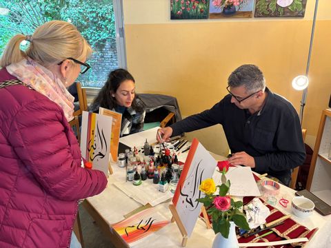 Ein Mann malt arabische Kaligraphie zwei Frauen beobachten den Künstler am Tisch