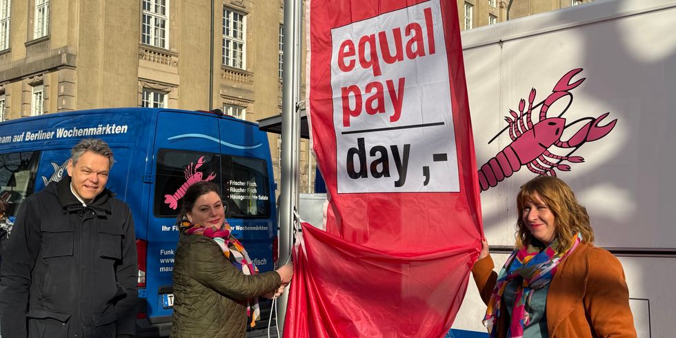 Ein Mann und zwei Frauen neben einer roten Flagge mit der Aufschrift "Equal Pay Day"