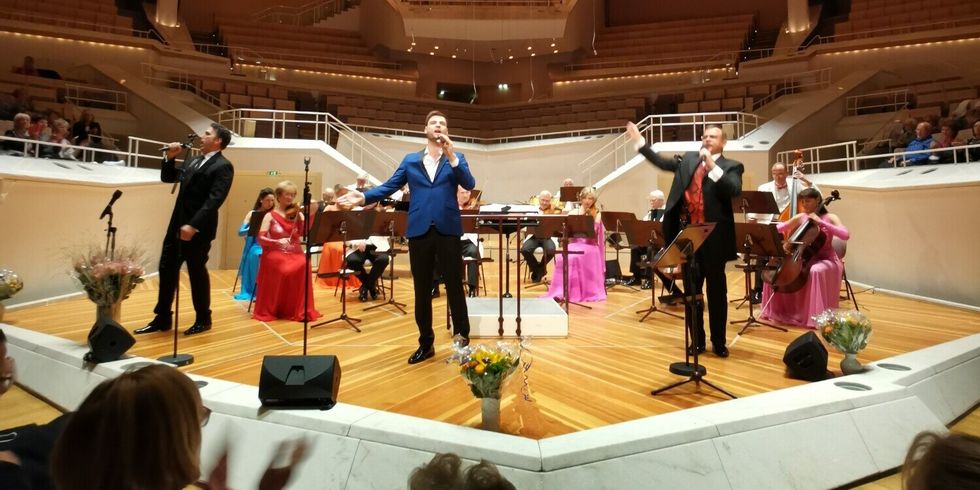 Auf einer Bühne in einem großen Saal machen Sänger und Musikanten zusammen Musik.