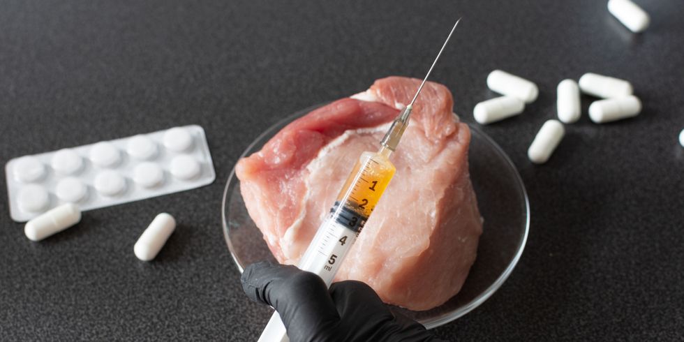 Hand mit einer Spritze vor einer Scheibe Fleisch, Tabletten liegen neben dem Fleisch