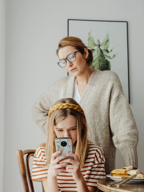 Eine Frau mit heller Strickjacke steht hinter einem Mädchen, das auf einem Stuhl sitzt. Das Mädchen trägt ein gestreiftes T-Shirt und hält ein Smartphone in beiden Händen. Auf dem Tisch liegen ein Teller mit Bananenscheiben und ein Glas. Im Hintergrund hängt ein gerahmtes Bild mit grünen Blättern an der Wand.