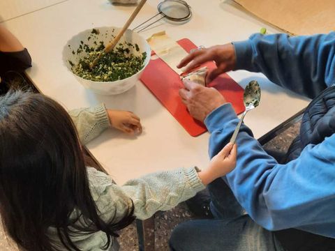 Ein Mädchen und eine erwachsene Person stellen an einem Tisch Blätterteigtaschen her, auf dem Tisch steht eine Schüssel mit Spinatfüllung