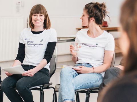 Zwei Frauen aus dem Projektteam der Berliner Ernährungscoaches sitzen im Stuhlkreis mit anderen Personen. Eine davon strahlt in die Kamera.