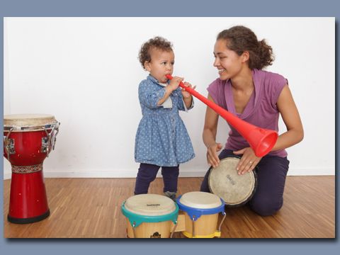 Frau kniet neben einem Kind, welches eine Kinderposaune bläst