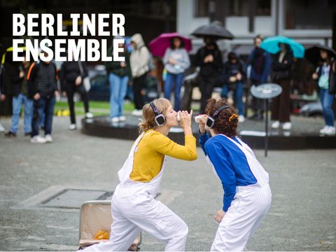 Zwei Frauen führen etwas auf einem Platz vor, im Hintergrund Publikum mit Regenschirmen