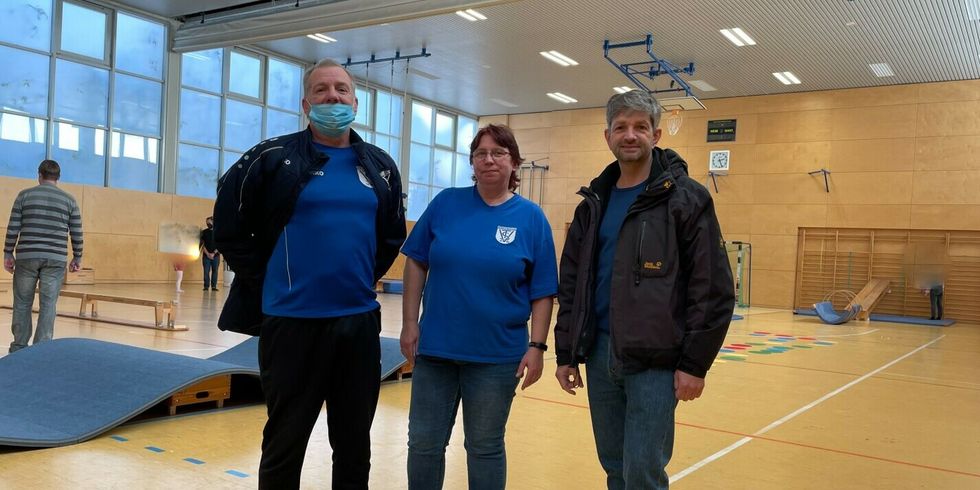 Drei Personen stehen zusammen in einer Turnhalle mit aufgebauten Spiel- und Sportgeräten.