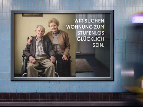 Auf dem Bild ist eine Werbetafel in einer U-Bahn-Station zu sehen. Die Tafel zeigt ein älteres Paar, wobei der Mann im Rollstuhl sitzt. Der Text auf der Tafel lautet: “WIR SUCHEN WOHNUNG ZUM STUFENLOS GLÜCKLICH SEIN.” Im Hintergrund bewegt sich ein Zug.
