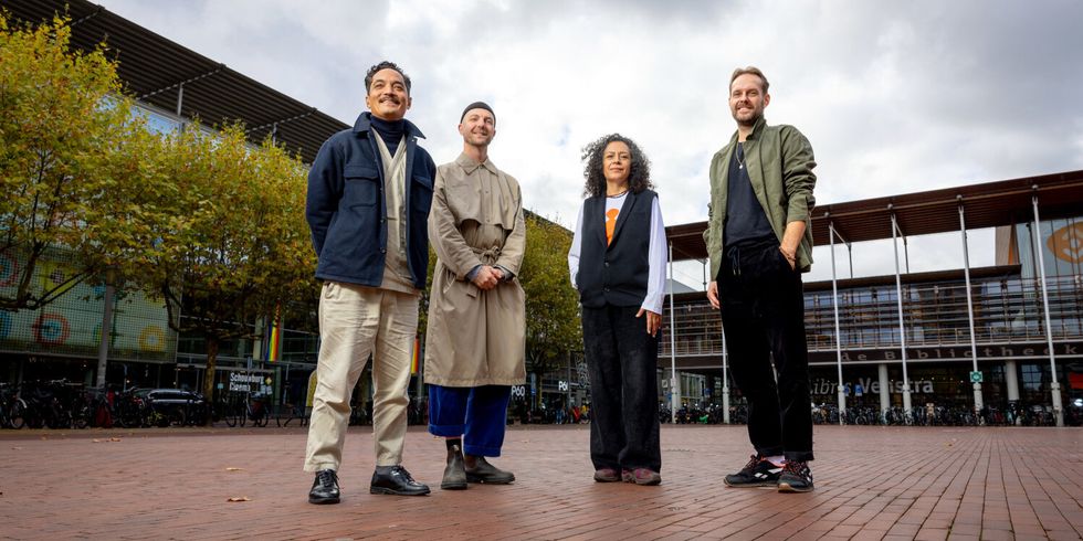 Vier Menschen stehen auf einem gefpflasterten Platz vor Bäumen und Gebäuden unter bewölktem Himmel