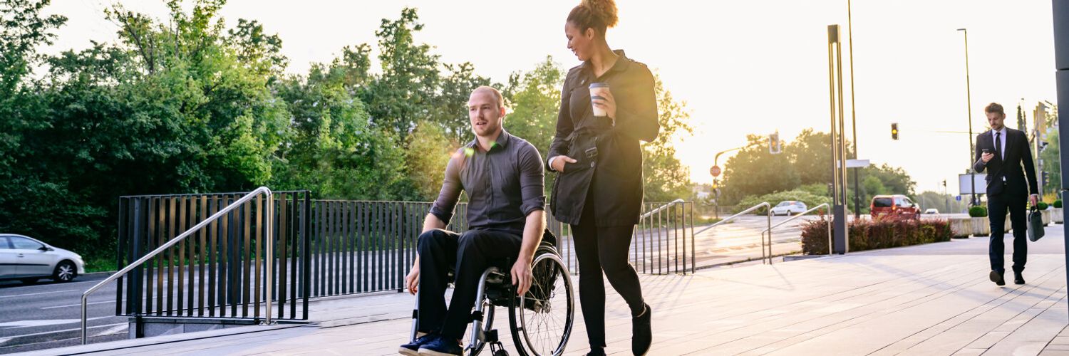 Zwei Personen, davon eine im Rollstuh sitzend unterhalten sich draußen auf einem Gehweg.