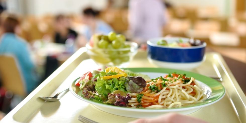 Person in einer Kantine hält ein Tablett mit Spaghetti und Tomatensauce, Salat und Weintrauben