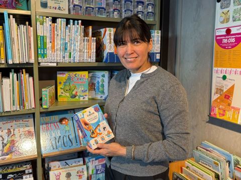 Mohidil Shamshidinova steht in der Else-Ury-Bibliothek vor einem Regal mit zahlreichen Kinderbüchern. Sie hält ein großformatiges Buch mit grünem Einband in den Händen und blickt darauf. Im Hintergrund sind weitere Bücherregale mit bunten Buchrücken zu sehen. Rechts an der Wand hängen mehrere Plakate und Informationsblätter, die farbig gestaltet sind. Links im Bild befinden sich weitere Bücherregale mit roten und weißen Buchrücken. Die Szene wirkt hell und freundlich.
