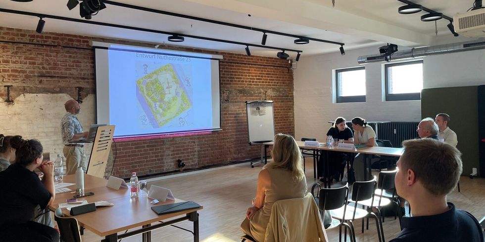 In einem Raum steht ein Mann neben einer Leinwand, auf die eine Präsentation projiziert wird. Im Raum sitzen mehrere Menschen und schauen zu ihm und der Leinwand.