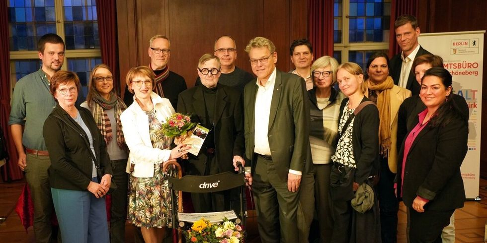 Eine Gruppe Menschen posiert für ein Gruppenfoto. Ein Mann in der Mitte hat Blumenstrauß und Urkunde.