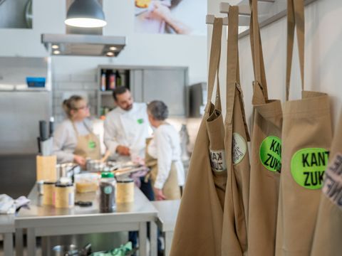 Mehrer beige Kochschürzen mit dem Logo der Kantine Zukunft hängen an einer Wand, im Hintergrund stehe drei Menschen in einer Küche