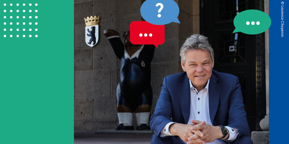 Ein Mann sitzt auf einer Treppe vor einem Gebäude. Hinter ihm ist ein Berliner Buddy Bär zu sehen und das Berliner Wappen an einer Wand. Von dem Mann ausgehend sind bunte Sprechblasen eingefügt.