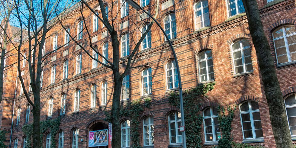 Hinter einer Reihe Bäume ist die mit Efeu bewachsene Fassade einen großen Backstein-Gebäudes.