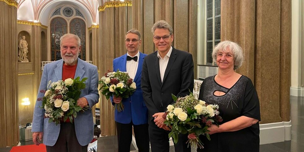 Vier Personen stehen in einer festlichen Halle. Drei Personen halten jeweils einen Blumenstrauß in der Hand.