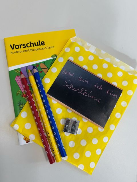 Ein gelbes Heft mit der Aufschrift 'Vorschule' liegt auf einem Tisch. Daneben befinden sich drei Buntstifte, ein Doppelspitzer aus Metall und eine kleine Tafel mit weißem Rahmen und gelbem Punktmuster. Auf der Tafel steht in weißer Schrift: 'bald bin ich ein Schulkind'.