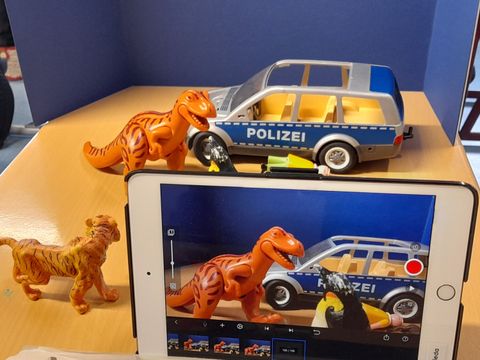 Tablet mit Aufnahme einer Soielzeug-Dino-Attacke auf ein Spielzeug-Polizeiauto
