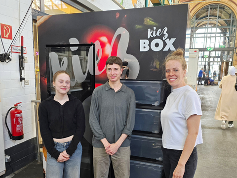Zwei Mitarbeitende der Markthalle IX und eine Mitarbeiterin der Verbraucherzentrale Berlin stehen vor der Kiez-Box in der Markthalle IX