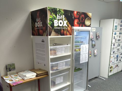 Kühlschrank mit der Aufschrift "Kiez Box", daneben ein Tisch mit Infomaterialien