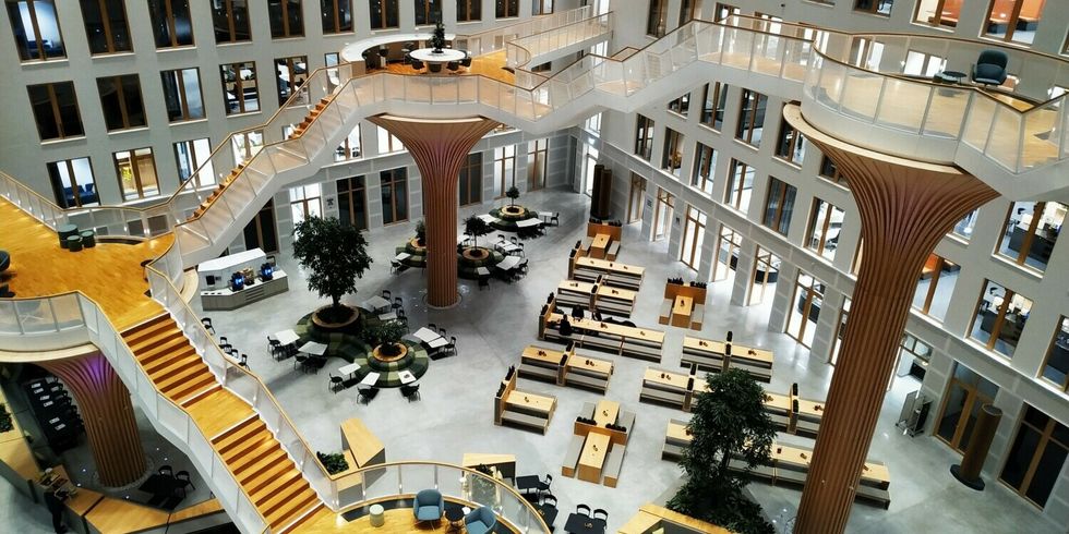 Eine große Halle. Auf dem Boden sind mehrere Tische, Bänke und auch Bäume. Treppen ziehen sich auf Säulen durch die Halle.