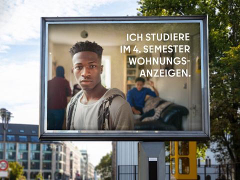 Auf einer Werbetafel in Berlin ist ein junger Mann abgebildet, der direkt in die Kamera blickt. Hinter ihm sind weitere Personen in einem Raum. Der Text auf der Tafel lautet: “ICH STUDIERE IM 4. SEMESTER WOHNUNGSANZEIGEN.” Im Hintergrund sieht man städtische Gebäude und Straßen.