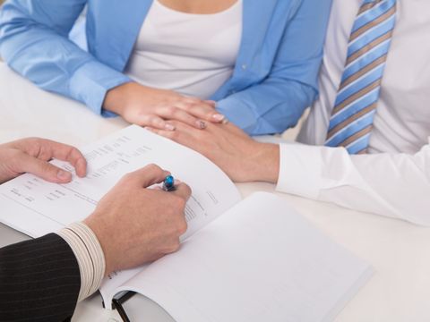 Mann im Anzug zeigt mit einem Stift auf ein Schriftstück, ihm gegenüber sitzt ein Hände haltendes Paar