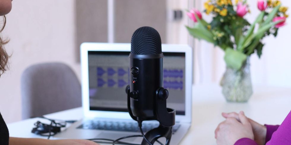 Zwei Frauen sitzen vor einem Mikrofon und im Hintergrund befinden sich ein Laptop und ein Blumenstrauß auf dem Tisch.