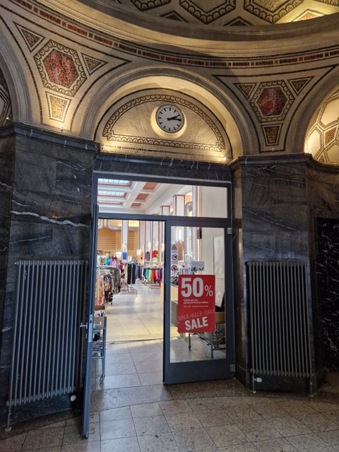 Glas-Eingangstür zu einem Bekleidungsladen Laden mit 50% Sale Poster an einer Glaswand und Uhr über dem Eingang in einer Marmorverzierten Halle mit Kuppel