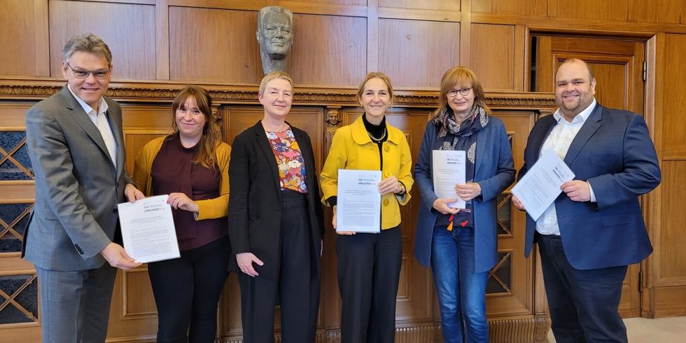 Vier Frauen und zwei Männer stehen zusammen in einem Raum und halten Kooperationsvereinbarungen in der Hand.