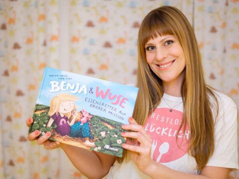 Wenke Heuts lächelt in die Kamera und hält das Kinderbuch Benja & Wuse Essensretter auf großer Mission in den Händen