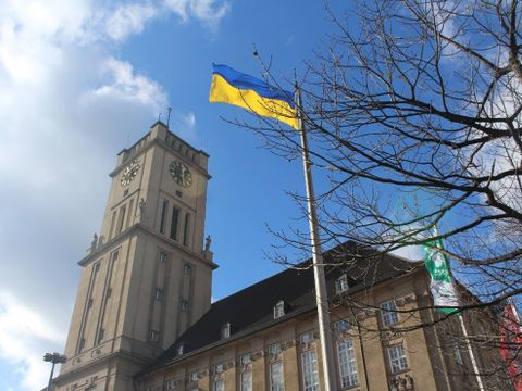 Vor einem großen Gebäude mit Uhrenturm ist an einem Fahnenmast eine gelb-blaue Fahne, die ukrainische Flagge, gehisst.