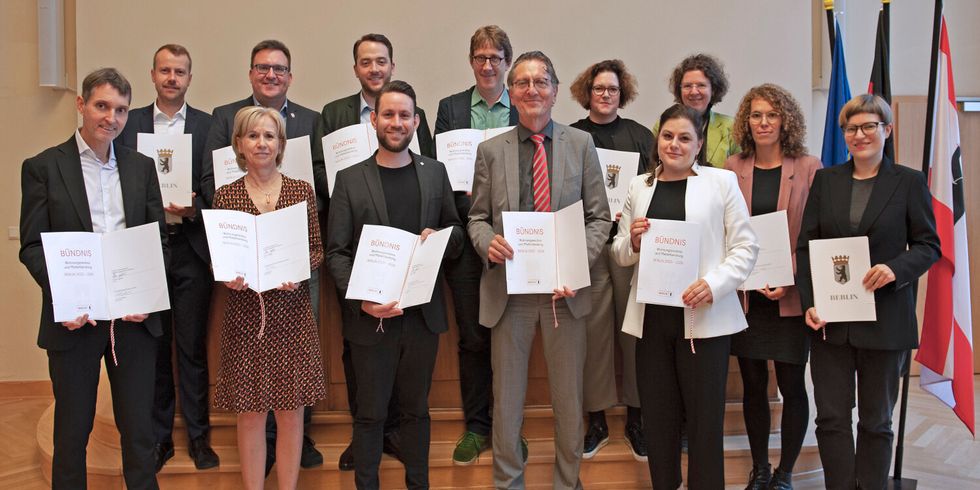 Dreizehn Personen mit unterschriebenen Bündnissen stehen zusammen in einem Raum.