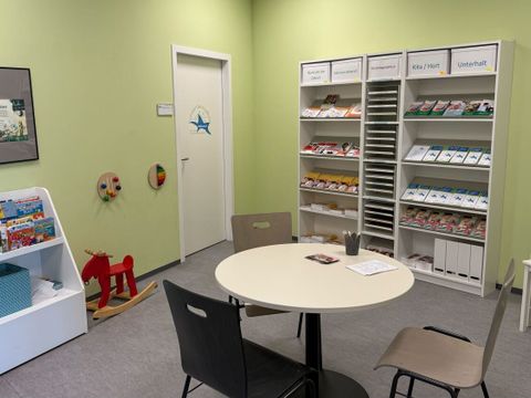 Im Wartebereich des FamilienServiceBüros Friedrichshain-Kreuzberg stehen ein runder Tisch mit drei Stühlen und eine kleine Kinderspielecke mit einem weißen Tisch und zwei Kinderstühlen. An der Wand befinden sich mehrere Regale mit sortierten Broschüren und Informationsmaterialien, die nach Themen wie 'Unterhalt' und 'Elterngeld' beschriftet sind. Links steht ein weißes Regal mit Kinderbüchern und Zeitschriften, daneben ein rotes Holzschaukelpferd und zwei an der Wand befestigte Holzspielzeuge. Die Wände sind hellgrün gestrichen. Der Raum wirkt aufgeräumt und freundlich gestaltet.