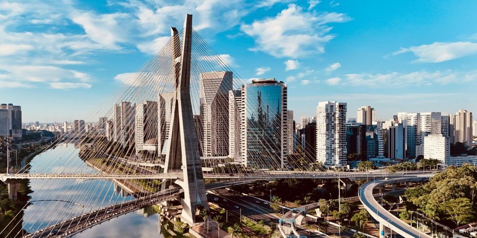 Foto von São Paulo am Tag. Im Vordergrund ist eine Brücke. Dahinter sieht man viele helle Hochhäuser der Stadt unter einem blauen Himmel.