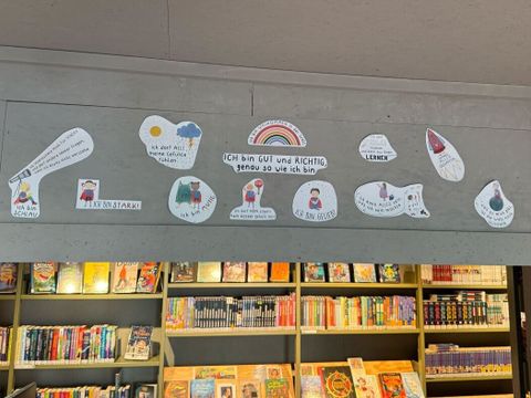 Über einem Bücherregal sind mehrere handgezeichnete Affirmationen auf weißen Papierformen angebracht. Jede Affirmation ist mit einer kleinen, farbigen Illustration versehen, darunter Regenbogen, Sonne, Herz und weitere Symbole. Die zentrale Affirmation steht in der Mitte und ist von einem Regenbogen umrahmt. Darunter sind weitere Affirmationen mit unterschiedlichen Motiven verteilt. Das Bücherregal ist gut gefüllt und zeigt zahlreiche bunte Buchrücken.