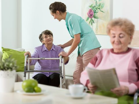 Im Aufenthaltsraum einer Pflegeeinrichtung sitzt eine Frau mit einer Zeitung an einem Tisch. Vor sich eine Kaffeetasse. Im Hintergrund hilft eine Pflegerin einer weiteren Frau mit einer Gehhilfe beim Aufstehen.