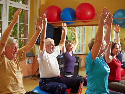 Eine Gruppe von Menschen verschiedenen Alters sitzen auf Gymnastikbällen und strecken die Hände in die Höhe.
