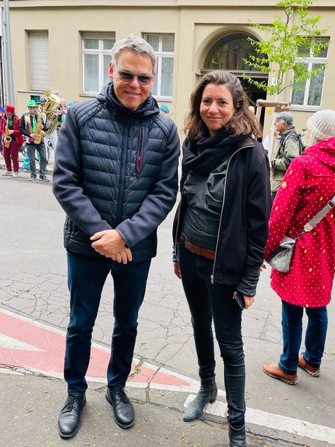 Ein Mann und eine Frau stehen auf einer Straße. Im Hintergrund sind bunt gekleidete Musiker und weitere Menschen.