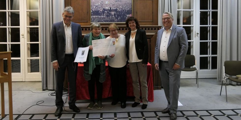Zwei Männer und drei Frauen stehen nebeneinander in einem Saal. Zwei Frauen halten zusammen einen Scheck in der Hand.