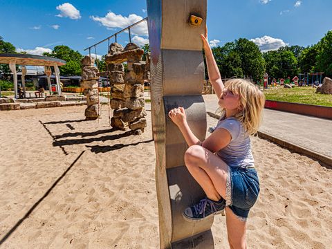Im odergrund sieht man ein Kletterelement, das aus mehreren aufeinander gestapelten großen, Sandsteinen besteht. Daran probiert sich gerade ein Mädchen hochzuziehen. Das Mädchen trägt eine blaue kurze Hose und ein graues T-Shirt. Sie hat blonde Haare. Im Hintergrund weitere Spielelemente. Auf dem Boden viel San.