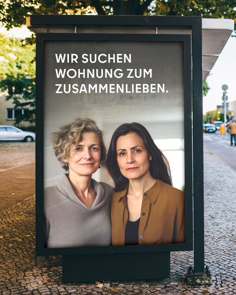 Das Bild zeigt eine Werbetafel an einer Straße. Auf der Tafel sind zwei Frauen abgebildet, die nebeneinander stehen und in die Kamera blicken. Der Text auf der Tafel lautet: “WIR SUCHEN WOHNUNG ZUM ZUSAMMENLIEBEN.” Im Hintergrund sind Gebäude, eine Straße und ein Fußgänger zu erkennen.