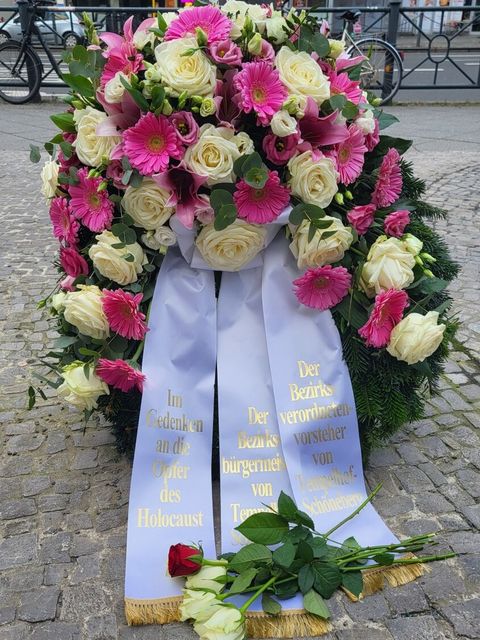 Ein rosa-weiß farbener Blumenkranz mit drei Kranzfahnen. 1. Fahne: Im Gedenken an die Opfer des Holocaust. 2. Fahne: Der Bezirksbürgermeister von Tempelhof-Schöneberg. 3. Fahne: Der Bezirksverordnetenvorsteher von Tempelhof-Schöneberg.