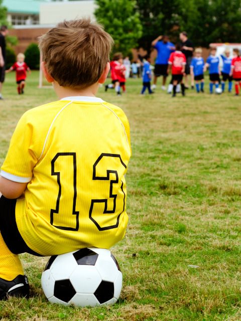 Ein kleiner Junge, auf einem Fußball sitzend, schaut zu einer Gruppe von Fußballern