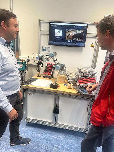 Zwei Männer stehen vor einem technischen Arbeitsplatz mit Roboterarm und Kleinteileboxen