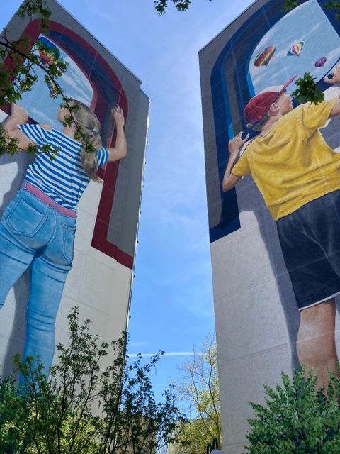 Zwei Hochhäuser mit Streetart: Bild von Mädchen und Junge, die jeweils durch ein Fenster auf einen Himmel mit Heißluftballons schauen.