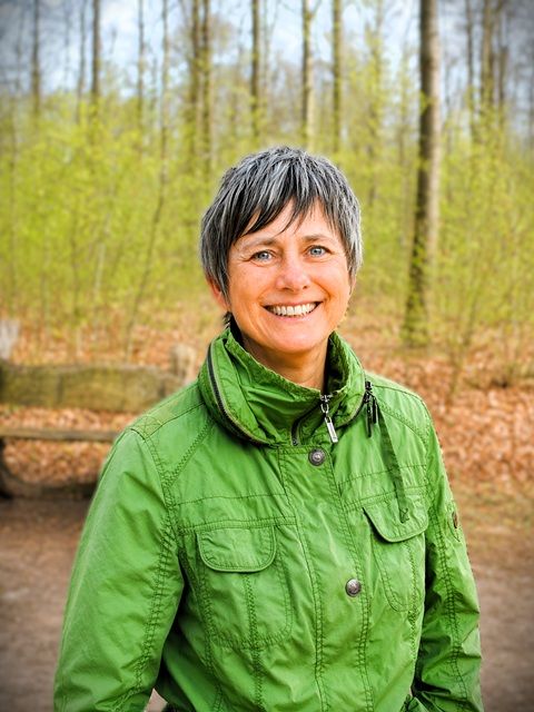 Anja Doms steht auf einem Waldweg und lacht den Betrachtenden zu. Sie trägt eine grasgrüne Jacke.
