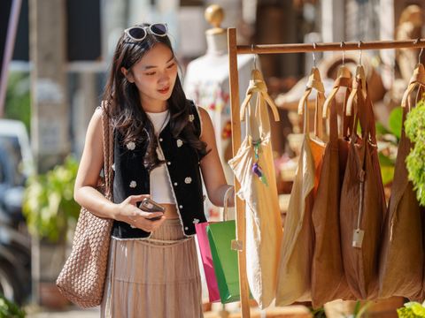Eine junge Frau mit Smartphone und Einkaufstüten betrachtet an einem Marktstand handgefertigten Taschen.