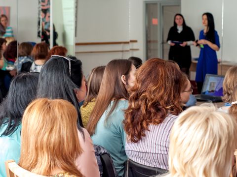 Publikum sitzt in einem Raum und hört zwei Frauen zu, die vorne stehend sprechen